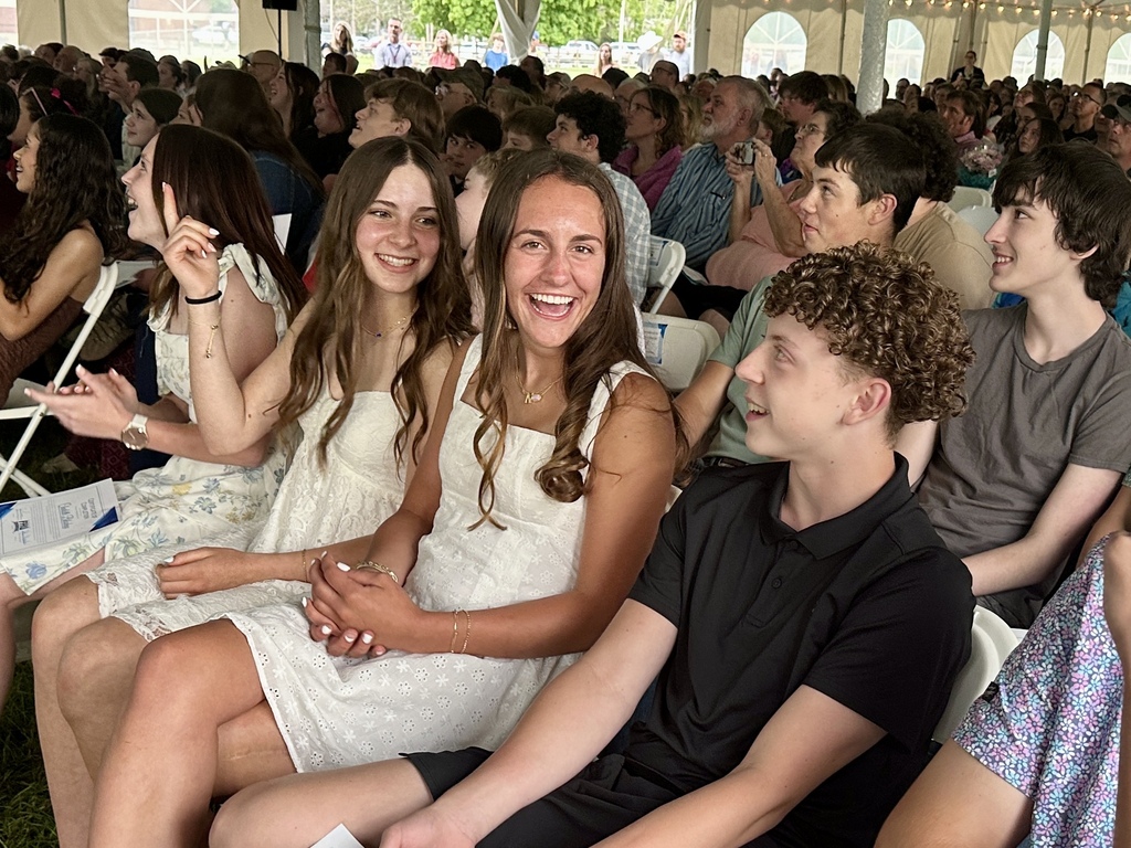 Students seated together in the audience smile and laugh during the celebration as families fill the tent behind them.