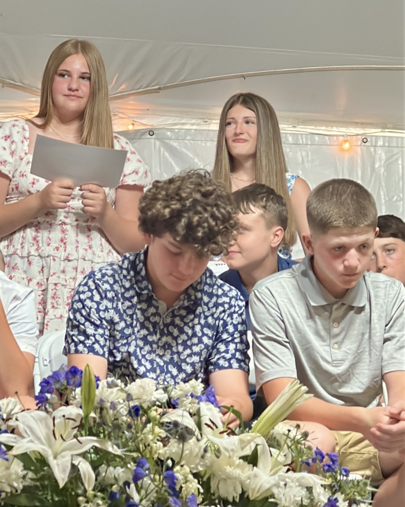 Several students participate in the ceremony while seated behind a large floral arrangement. Two students stand holding papers as they address the audience.