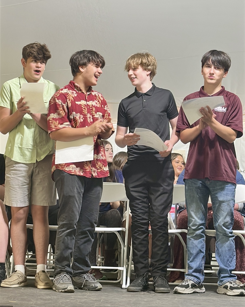 Four eighth-grade boys stand together on stage holding papers and smiling while participating in the celebration program.