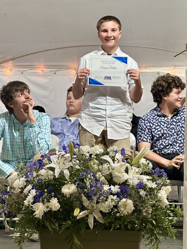 A smiling student stands on stage holding a certificate of completion while classmates sit behind him. A large blue-and-white floral arrangement is displayed in the foreground.