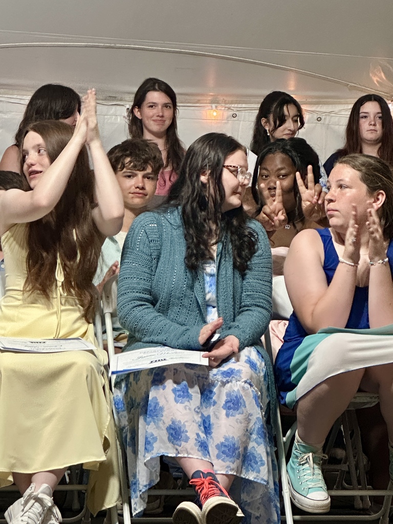 A group of eighth-grade students seated on stage applaud and smile during the program. One student in the background playfully flashes a peace sign toward the camera.