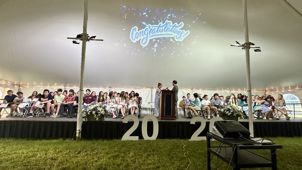 A wide stage view shows students seated on both sides of the podium beneath a projected "Congratulations!" graphic, with large white numbers spelling out 2026 in front of the stage.