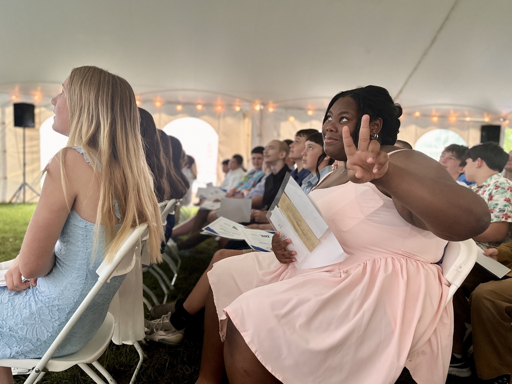 An eighth-grade student seated among classmates smiles and flashes a peace sign toward the camera while holding ceremony materials.