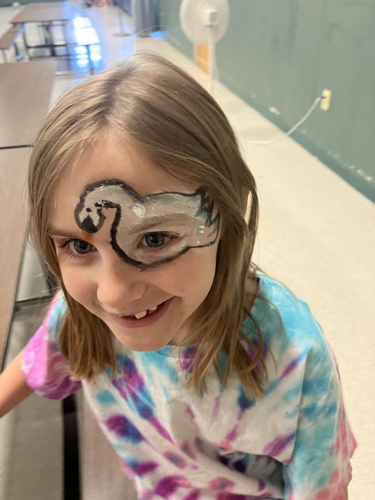 A young girl with a tie-dye shirt smiles up at the camera, showing off her face paint that features a detailed white swan design wrapping around her left eye.