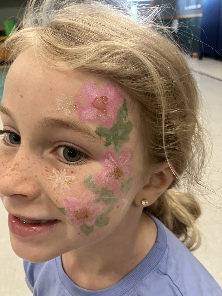 A close-up portrait of a young blonde girl smiling, featuring elaborate face paint of pink flowers and green leaves with gold glitter details on her left cheek and forehead.
