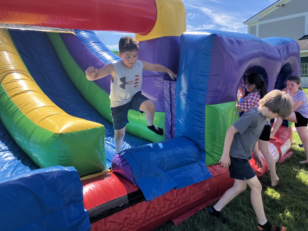 : A boy wearing a grey sleeveless Under Armour shirt jumps on a colorful, inflatable bounce house obstacle course outdoors on a sunny day, while other children navigate through a nearby tunnel section.