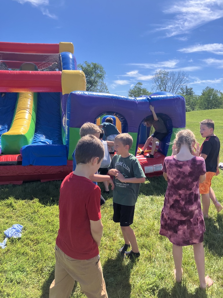 A group of children gather around a large, brightly colored inflatable bounce house structure on a grassy lawn under a blue sky with light clouds. Some children are talking on the grass, while others climb inside the inflatable tunnels. IMG_3479.jpg Alt Text: A group of children with various colorful face paint designs sit lined up against a dark green wall labeled "Cool Down Corner," holding basketballs, soccer balls, and footballs.