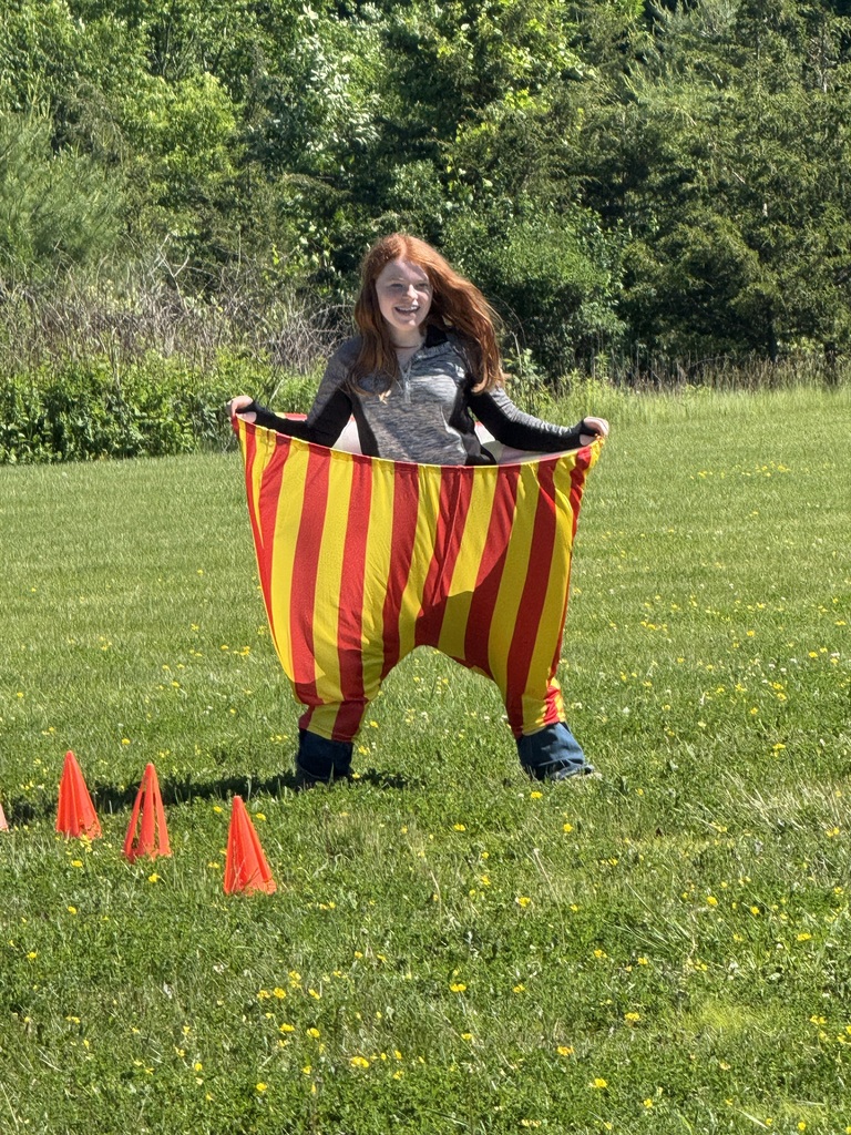 A smiling girl with long red hair stands in a grassy field holding out a pair of comically large, oversized red-and-yellow striped pants. A row of small orange cones is placed on the grass in front of her.
