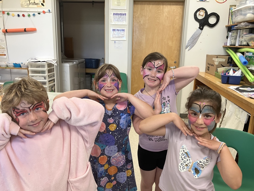 : Four young children stand together in a classroom, each posing with their hands under their chins to showcase their matching butterfly face paint designs in various colors. .