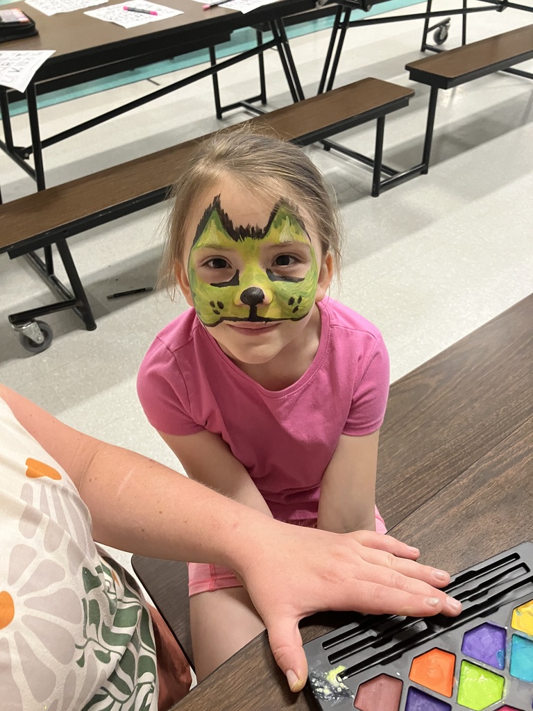 A young girl in a pink shirt sits at a school cafeteria table, looking directly at the camera with her face painted to look like a green and yellow cartoon animal. An adult's arm and a face paint palette are visible in the foreground.