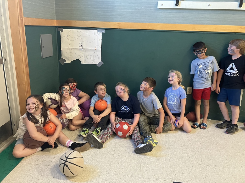 A group of children with various colorful face paint designs sit lined up against a dark green wall labeled "Cool Down Corner," holding basketballs, soccer balls, and footballs.