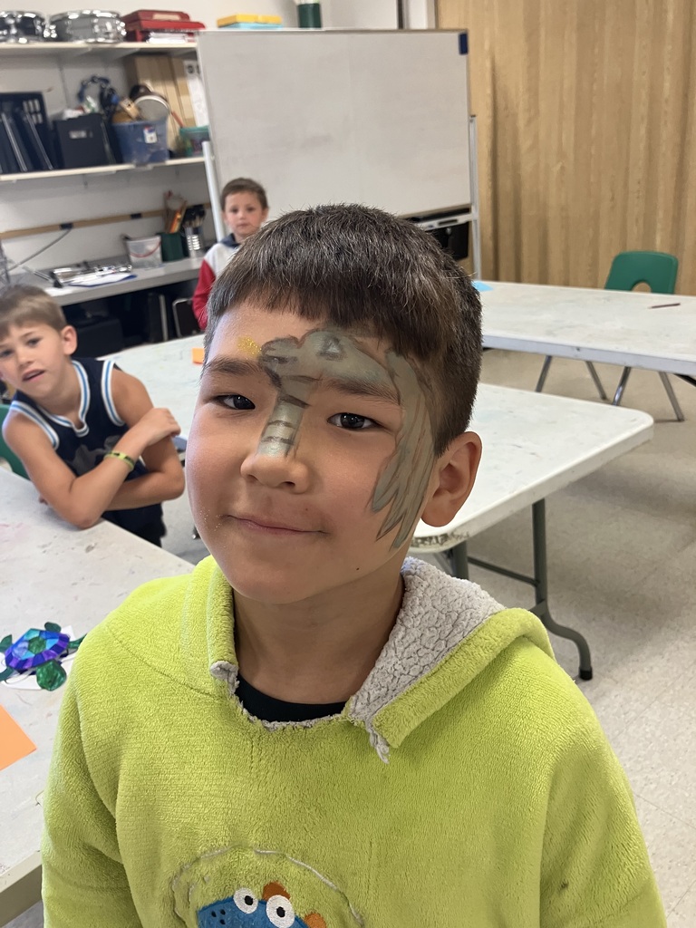 A young boy in a bright green hoodie smiles with a green lizard or dinosaur design painted across his forehead and down the side of his face, with other children visible in the classroom background.
