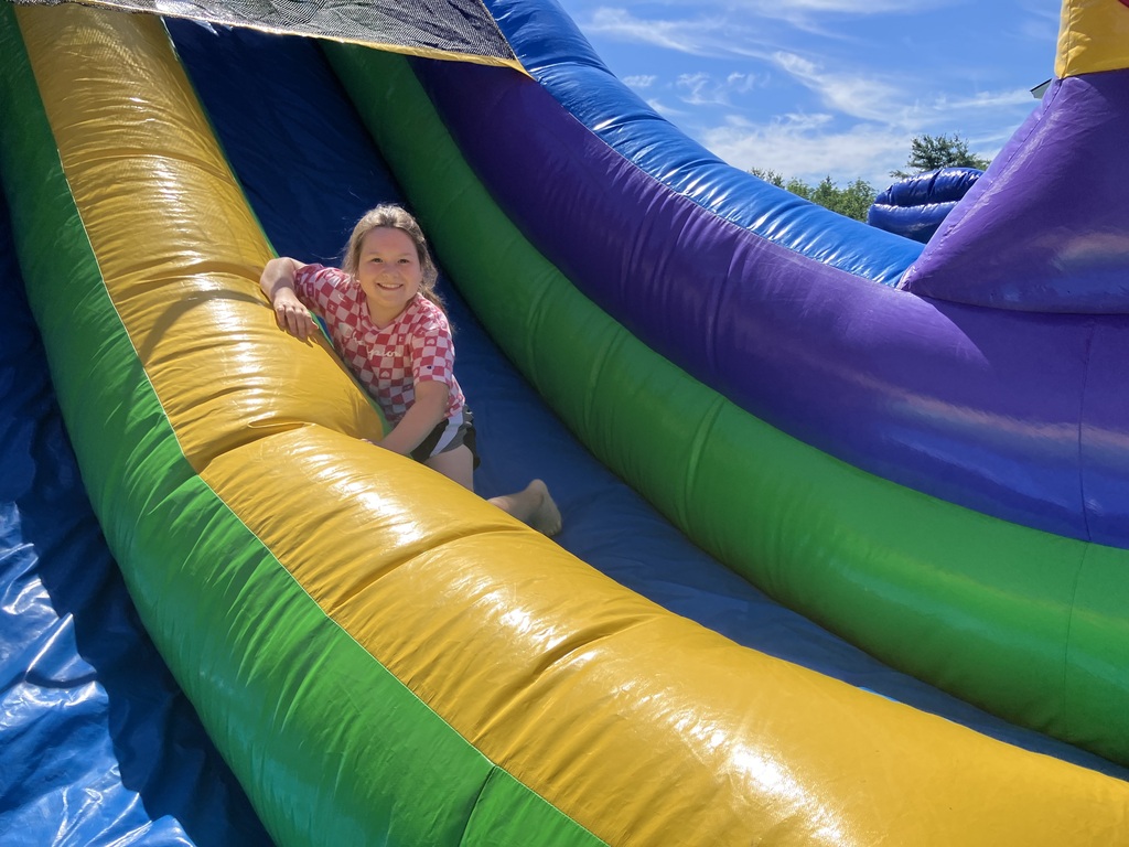 A young girl with a blonde braid, wearing a maroon tie-dye dress, sitting at the bottom of a large inflatable slide. She is smiling directly at the camera. The inflatable structure is made of bold panels of blue, green, yellow, purple, and red, sitting outdoors on green grass under a clear blue sky.