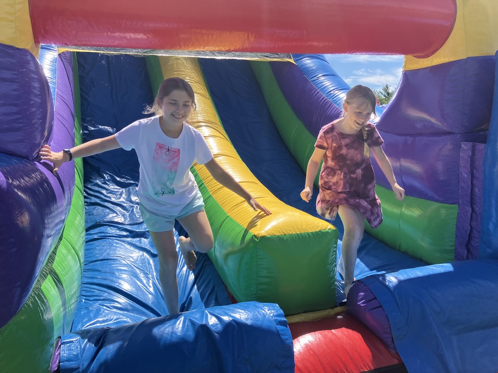 Two young girls smiling and playing inside a bright, multi-colored inflatable bounce house. The girl on the left is wearing a white graphic t-shirt and light blue shorts, balancing on one foot. The girl on the right is wearing a maroon tie-dye dress, caught mid-motion with a happy expression. The background features vibrant blue, green, yellow, and purple inflatable walls under a sunny sky.