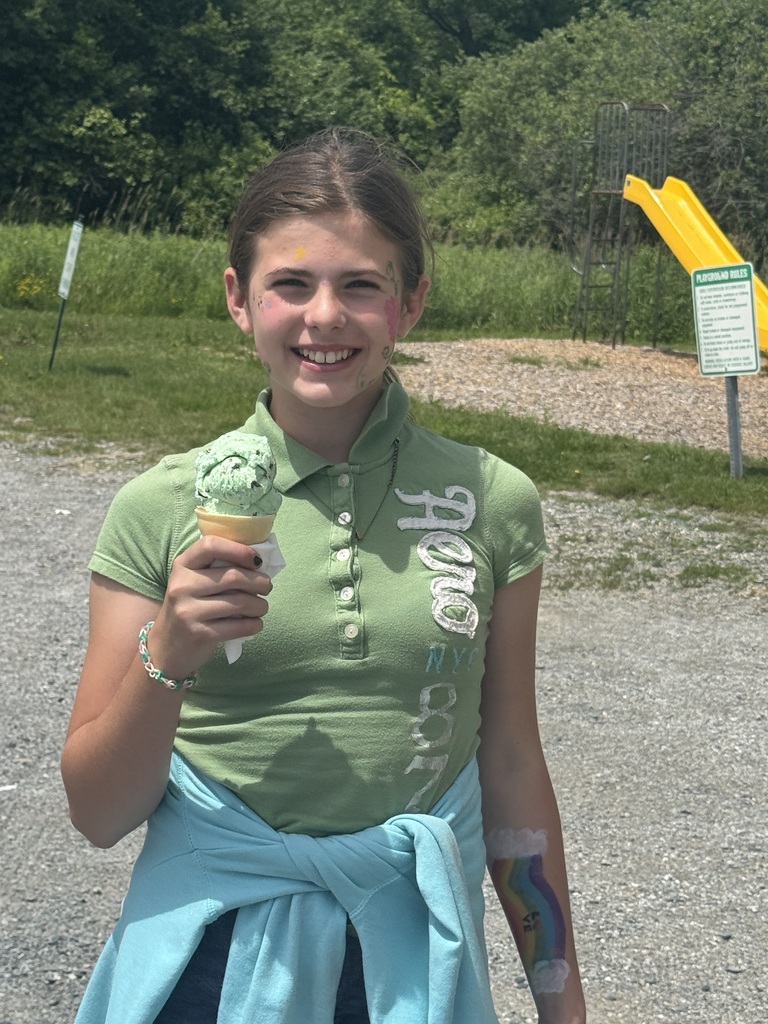 A smiling girl stands outdoors holding a waffle cone with green mint chocolate chip ice cream. She is wearing a green polo shirt and has a light blue sweatshirt tied around her waist. She has subtle face paint on her cheek and a colorful rainbow painted on her left forearm. A playground with a yellow slide is visible in the background.