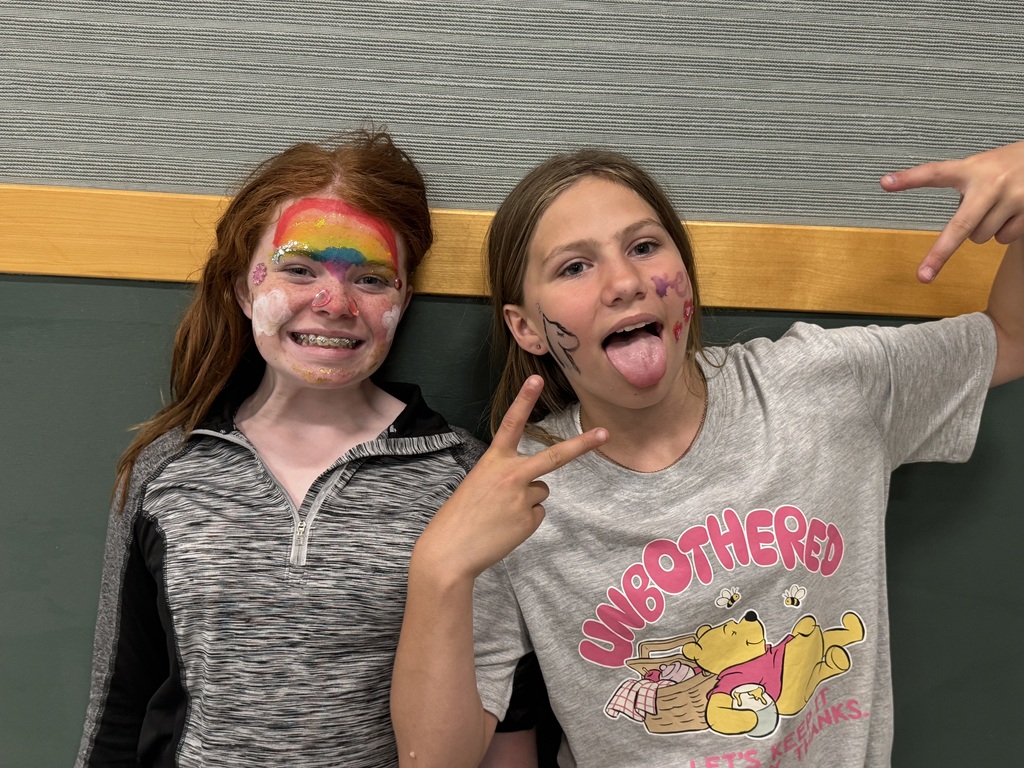 : Two girls pose playfully against an indoor wall with gray texturing and green paneling. The girl on the left has a vibrant rainbow painted across her forehead, white painted clouds on her cheeks, and smiles brightly with braces. The girl on the right sticks her tongue out, makes peace signs with both hands, has face paint on her cheek, and wears a gray t-shirt featuring Winnie the Pooh.