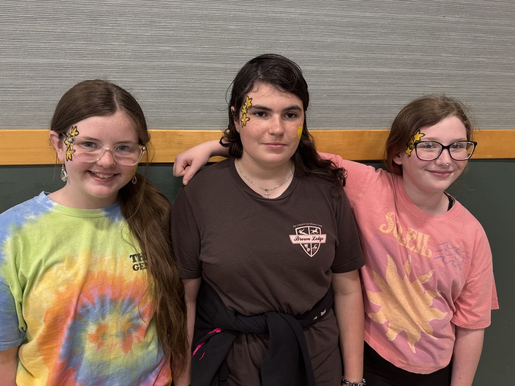 Three girls pose closely together in front of a wall with a gray textured upper half and a green lower half. All three have yellow star painted designs on the sides of their faces near their eyes. The girl on the left wears a colorful tie-dye t-shirt and glasses; the center girl wears a brown t-shirt; and the girl on the right wears a pink t-shirt with a sun graphic and glasses.