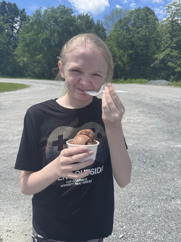 A young person stands outside on a gravel path, making a playful squinting expression while eating chocolate ice cream from a paper cup with a plastic spoon. They are wearing a black t-shirt with a video game controller graphic that reads "Game Outside." The background shows lush green trees under a partly cloudy sky.