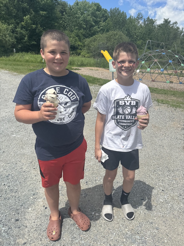 Two smiling boys stand next to each other on a gravel area under a bright, sunny sky. The boy on the left wears a dark blue "Cape Cod" t-shirt, red shorts, and brown Crocs, holding an ice cream cone. The boy on the right has red and black painted designs on his face, wears a white graphic t-shirt, black shorts, and gray slides, and is holding a pink ice cream cone. A playground with a yellow slide and dome climber is visible in the background.
