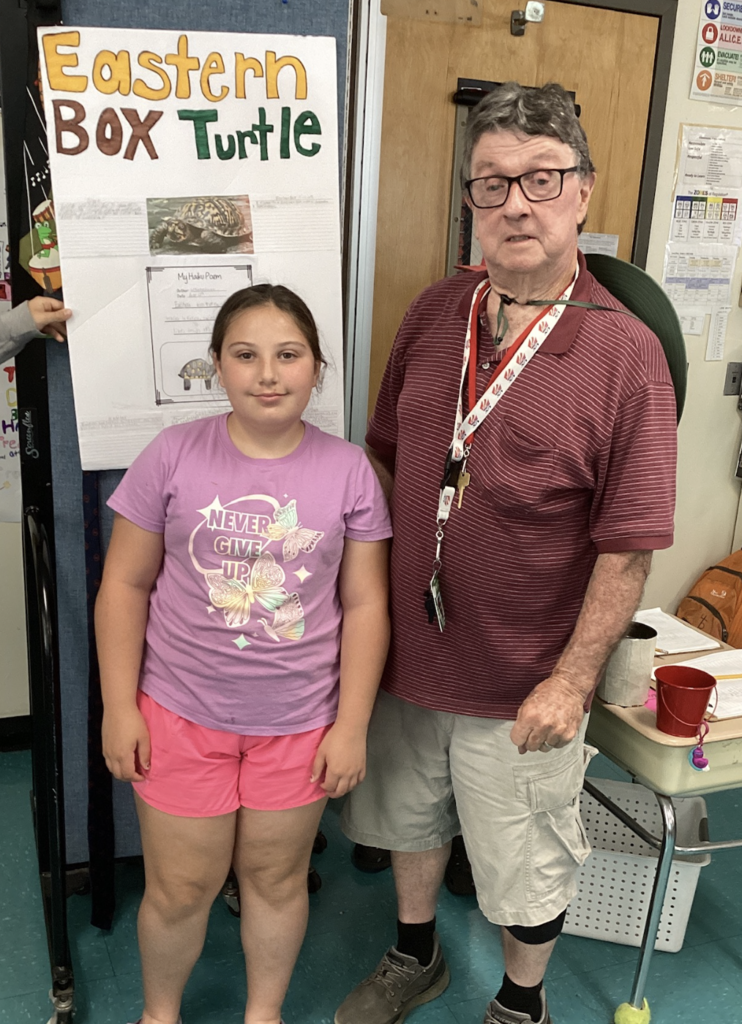 A student in a purple shirt and pink shorts stands with a guest presenter in front of a project board about the Eastern Box Turtle, featuring student writing and illustrations.