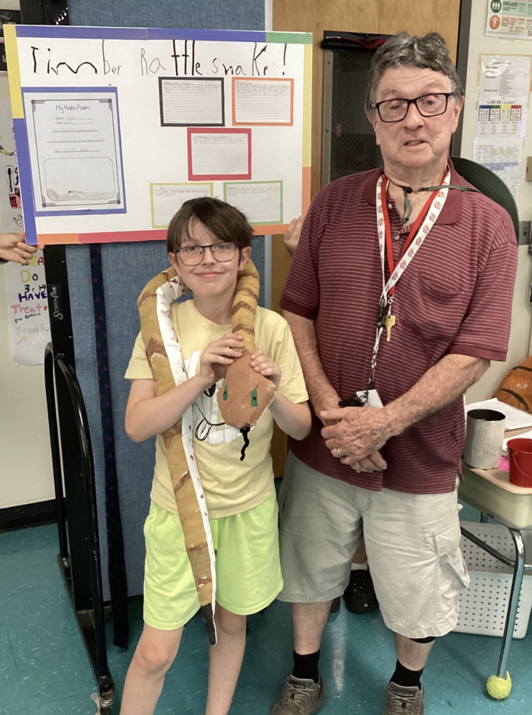 A student wearing glasses and neon green shorts stands beside a guest presenter while holding a handmade snake model. Behind them is a project board about the Timber Rattlesnake with student research and writing.