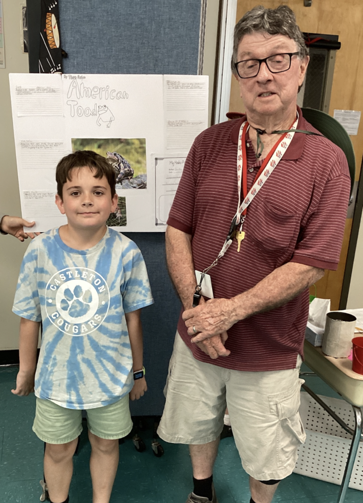 A student wearing a blue tie-dye Castleton Cougars shirt poses with a guest presenter in front of a wildlife research display about the American Toad.