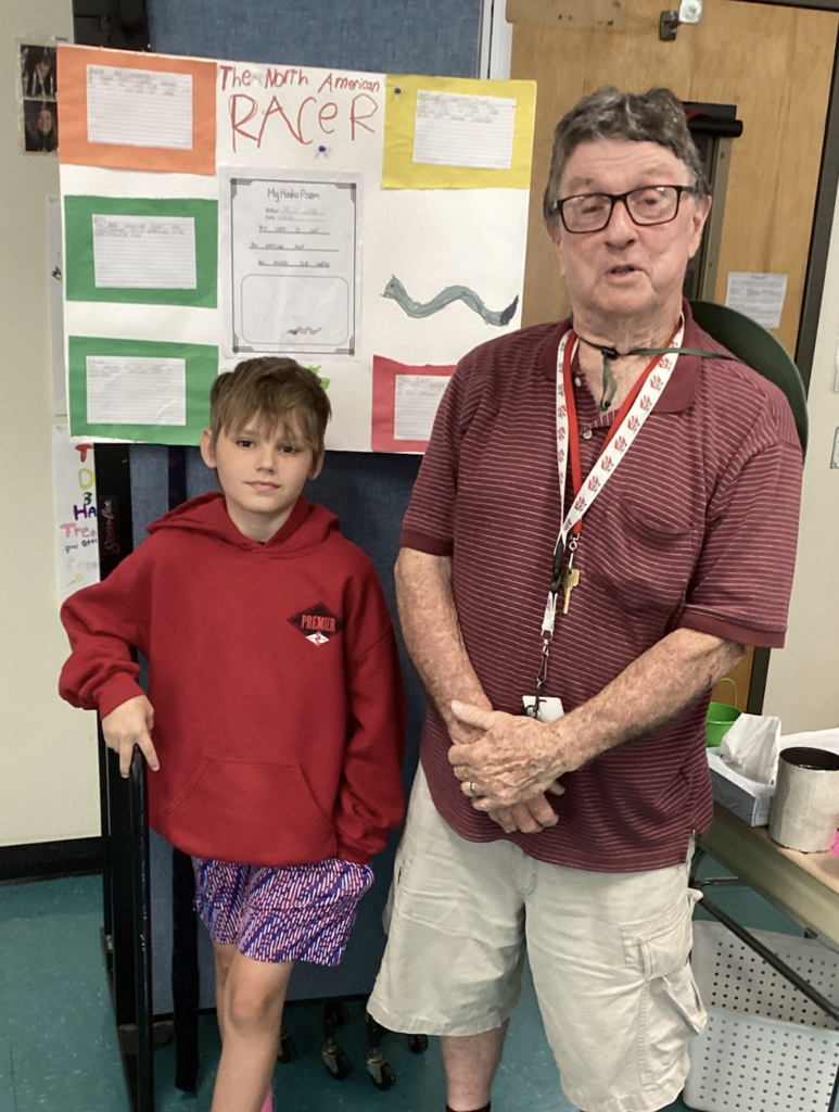 A student in a red hoodie stands beside a guest presenter in front of a colorful project board about the North American Racer snake, featuring student-written facts and artwork.
