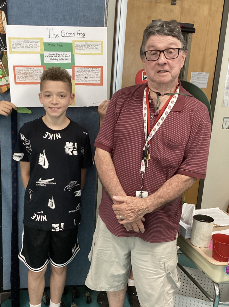 A student wearing a black Nike outfit poses with a guest presenter in front of a research display about the Green Frog. The board includes facts, a haiku poem, and student writing.