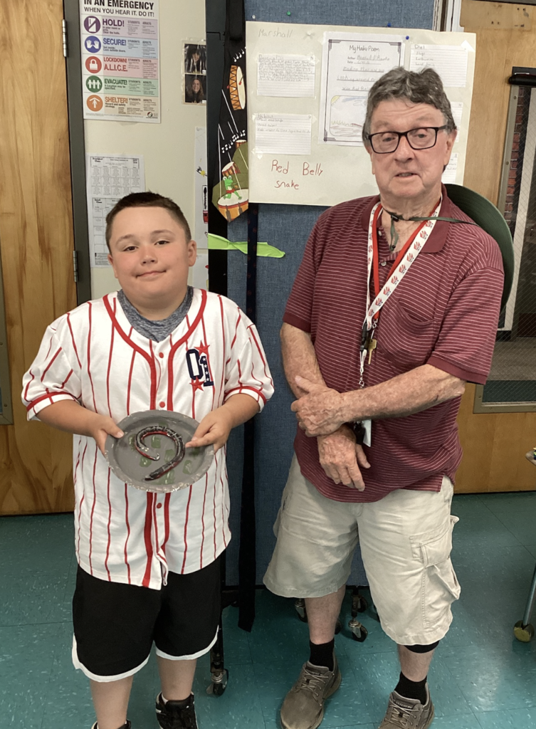 A student wearing a striped baseball jersey holds a model of a snake while standing beside a guest presenter. A research display about the Red-Bellied Snake is shown behind them.