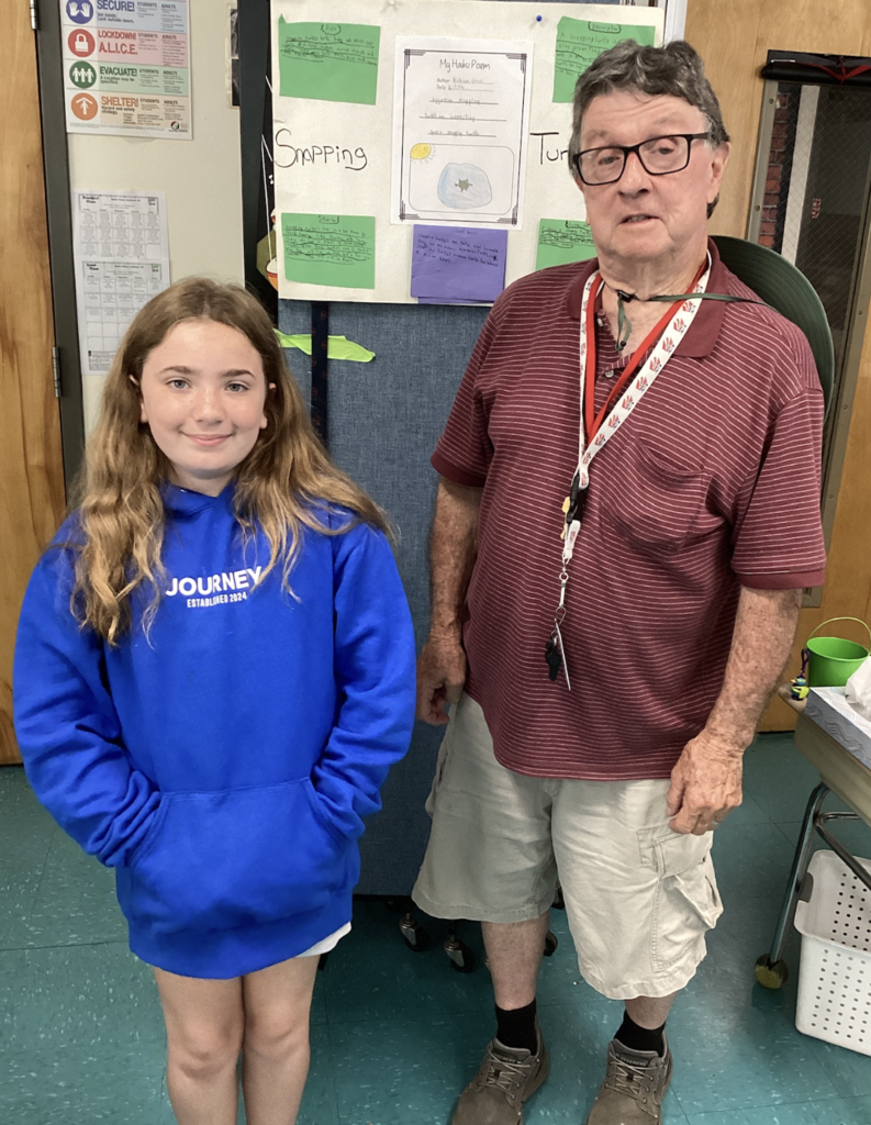 A student in a bright blue hoodie poses with a guest presenter next to a wildlife project display board featuring information about a snapping turtle.