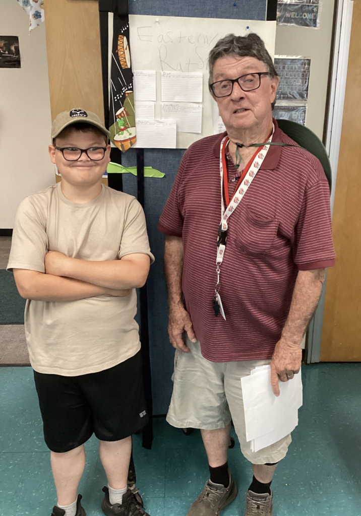A student wearing a tan shirt and baseball cap stands beside a guest presenter in a classroom during a wildlife research project presentation. A display board about the Eastern Rat Snake is visible in the background.