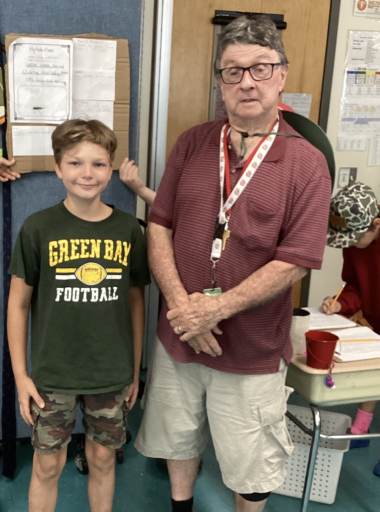 A student wearing a Green Bay Football shirt poses with a guest presenter next to a wildlife research display featuring a haiku poem and information about a local animal species.