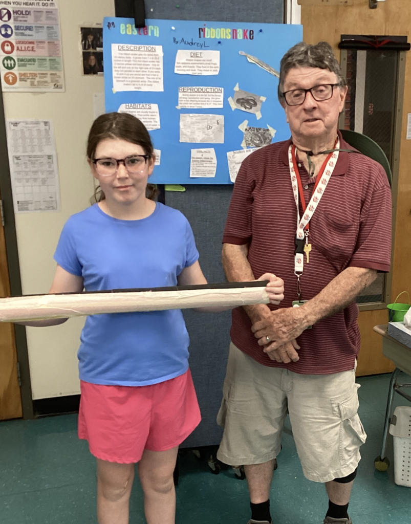 A student wearing glasses and a blue shirt holds a large handmade snake model while posing with a guest presenter. Behind them is a research display about the Eastern Ribbon Snake.