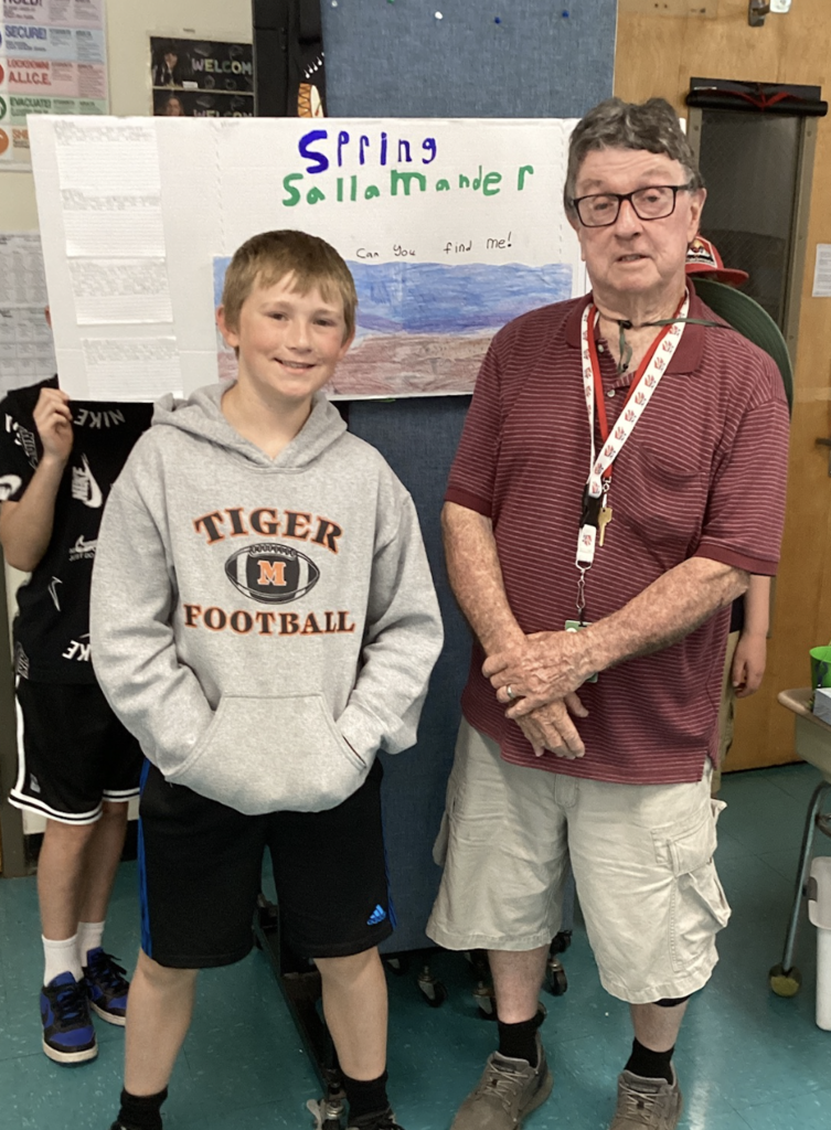 A student in a gray Tiger Football sweatshirt stands beside a guest presenter in front of a project board titled “Spring Salamander,” which includes student research and artwork.