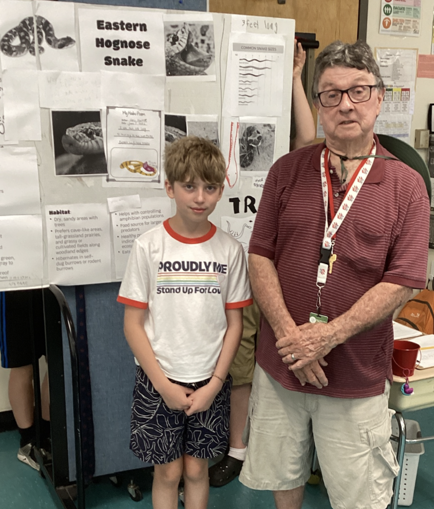A student wearing a white “Proudly Me” shirt stands with a guest presenter in front of a detailed research display about the Eastern Hognose Snake, including habitat information, photographs, and student work.