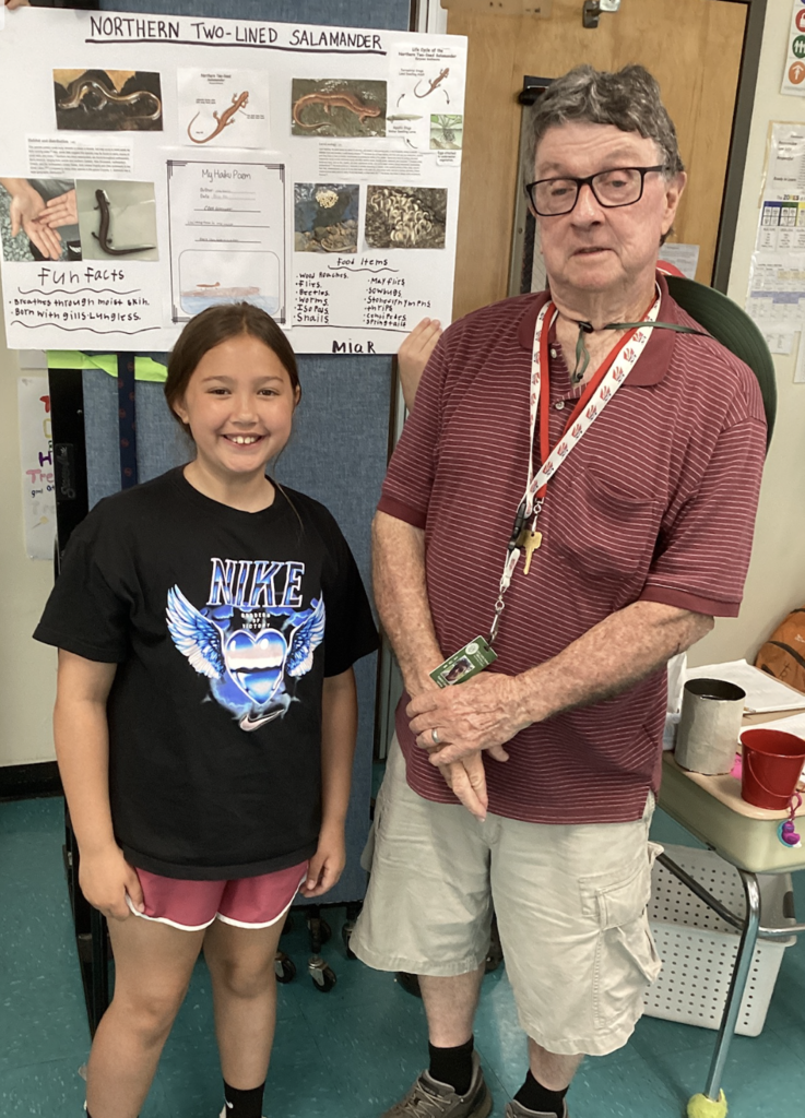 A smiling student stands beside a guest presenter in front of a research display about the Northern Two-Lined Salamander. The project board includes photos, facts, a life cycle diagram, and student writing.