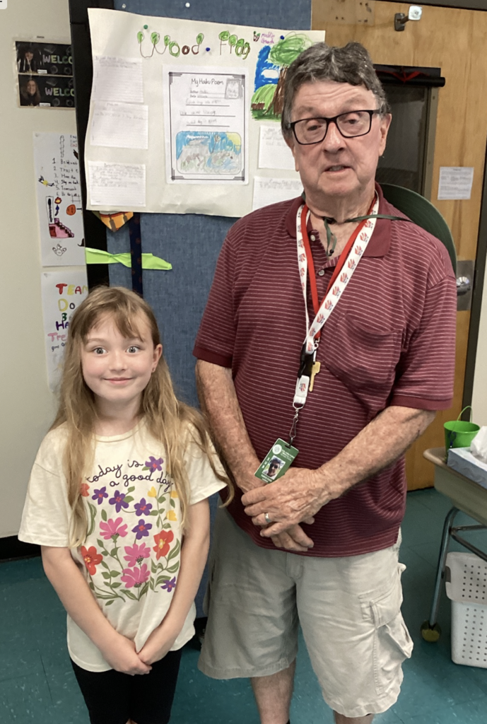 A student wearing a floral-themed shirt stands beside a guest presenter in front of a project board about the Wood Frog, featuring student writing, illustrations, and a haiku poem.