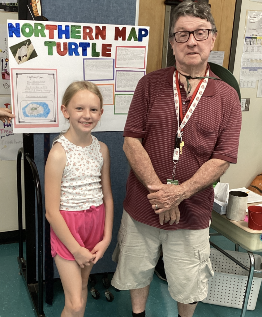 A smiling student stands with a guest presenter in front of a colorful research display about the Northern Map Turtle. The board includes facts, student writing, and a hand-drawn turtle illustration.