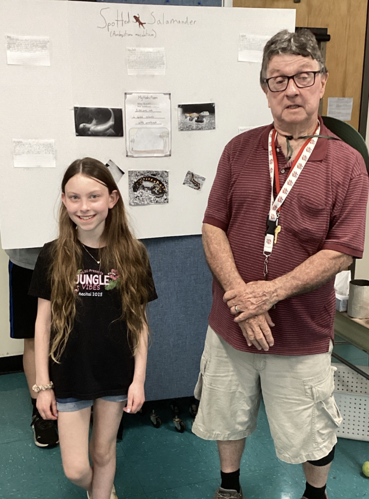 A student wearing a black “Jungle Vibes” shirt poses beside a guest presenter in front of a research board about the Spotted Salamander, featuring photos and student-written information.