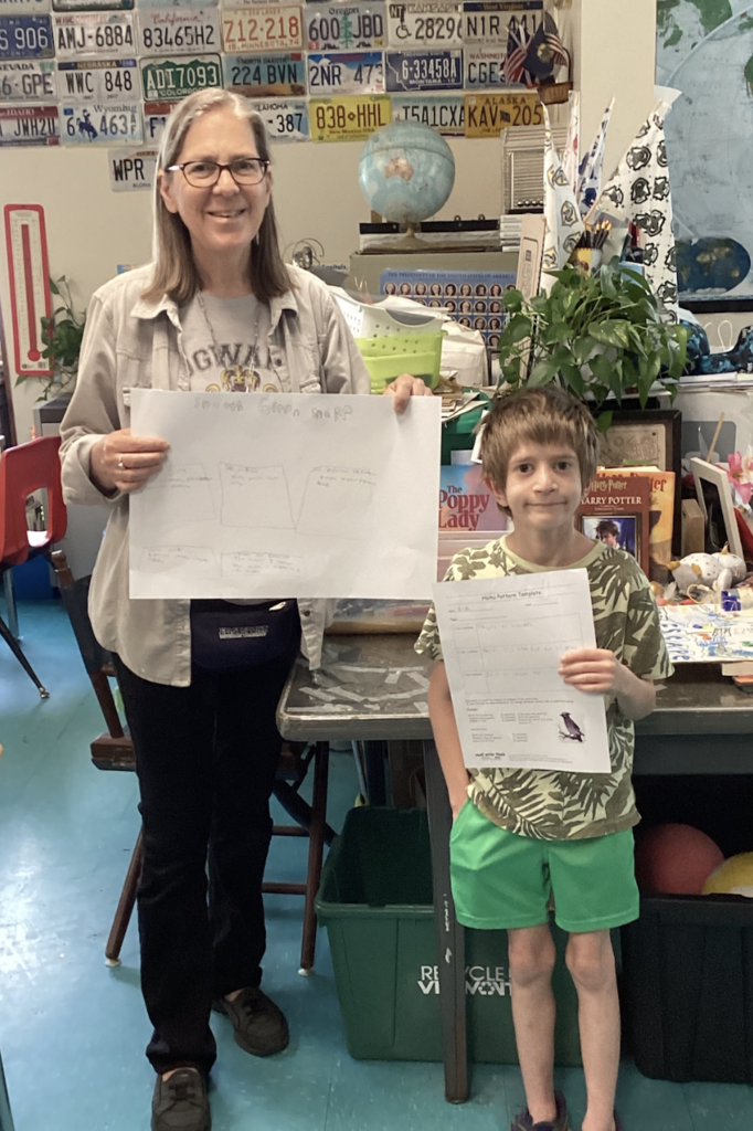 A student holds a wildlife research project and a completed worksheet while standing next to a teacher. The display focuses on a green snake and includes student research notes and illustrations.