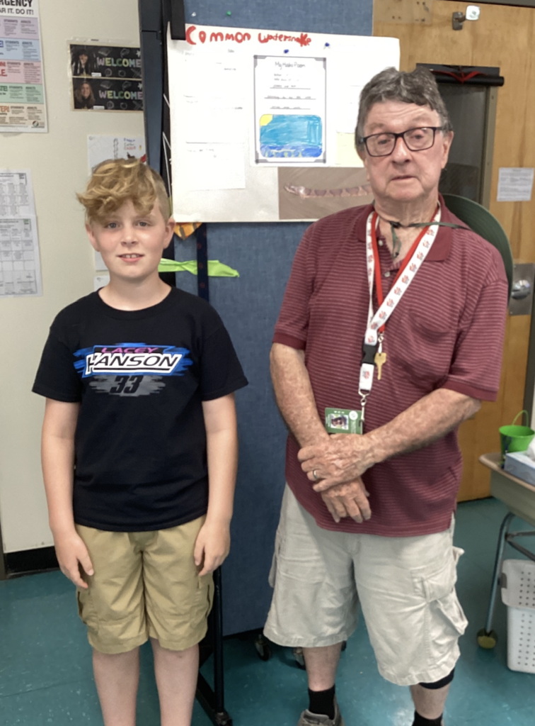A student stands beside a guest presenter in front of a research display about the Common Watersnake. The project board includes student writing, a haiku poem, and an illustration of a watersnake habitat.