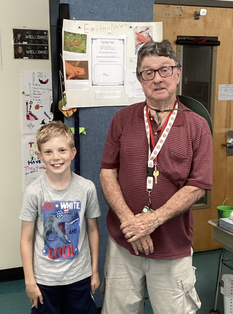 A smiling student stands beside a guest presenter in front of a project board about the Eastern Newt. The display includes photographs, a haiku poem, and information about the amphibian’s life cycle and habitat.
