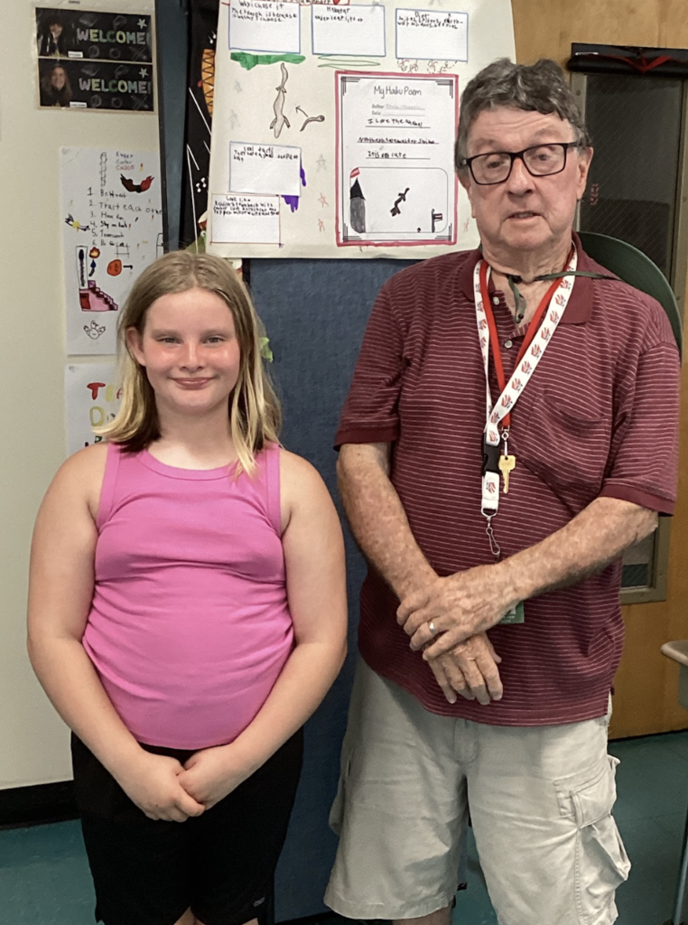 A student wearing a pink tank top poses with a guest presenter in front of a wildlife research project featuring a salamander life cycle illustration, a haiku poem, and student-written facts.