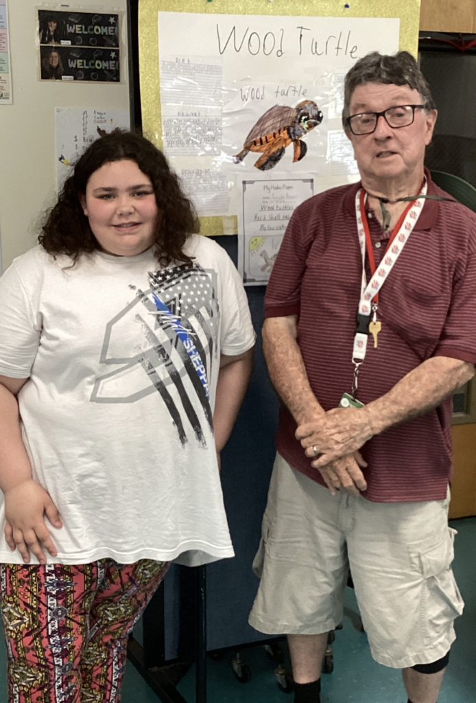 A student stands beside a guest presenter in front of a research project about the Wood Turtle. The display features student writing, drawings, and information about the turtle’s habitat and characteristics.
