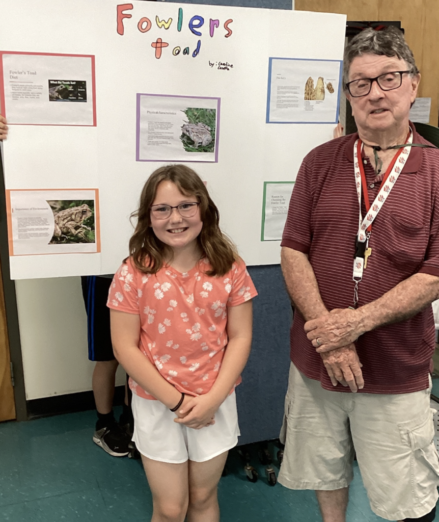 A smiling student stands in front of a large research board about Fowler’s Toad while posing with a guest presenter. The display includes information about diet, physical characteristics, and environmental importance.