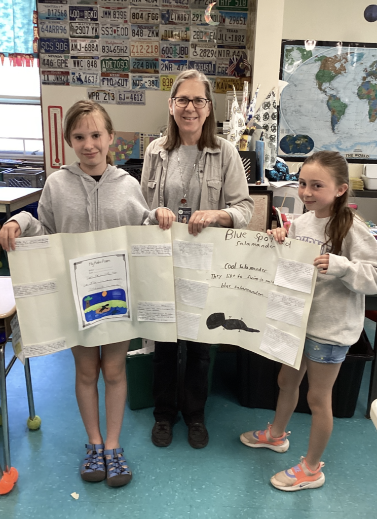 Two students stand with their teacher while displaying a large research board about the Blue-Spotted Salamander. The project includes student writing, a haiku poem, and a hand-drawn salamander illustration.