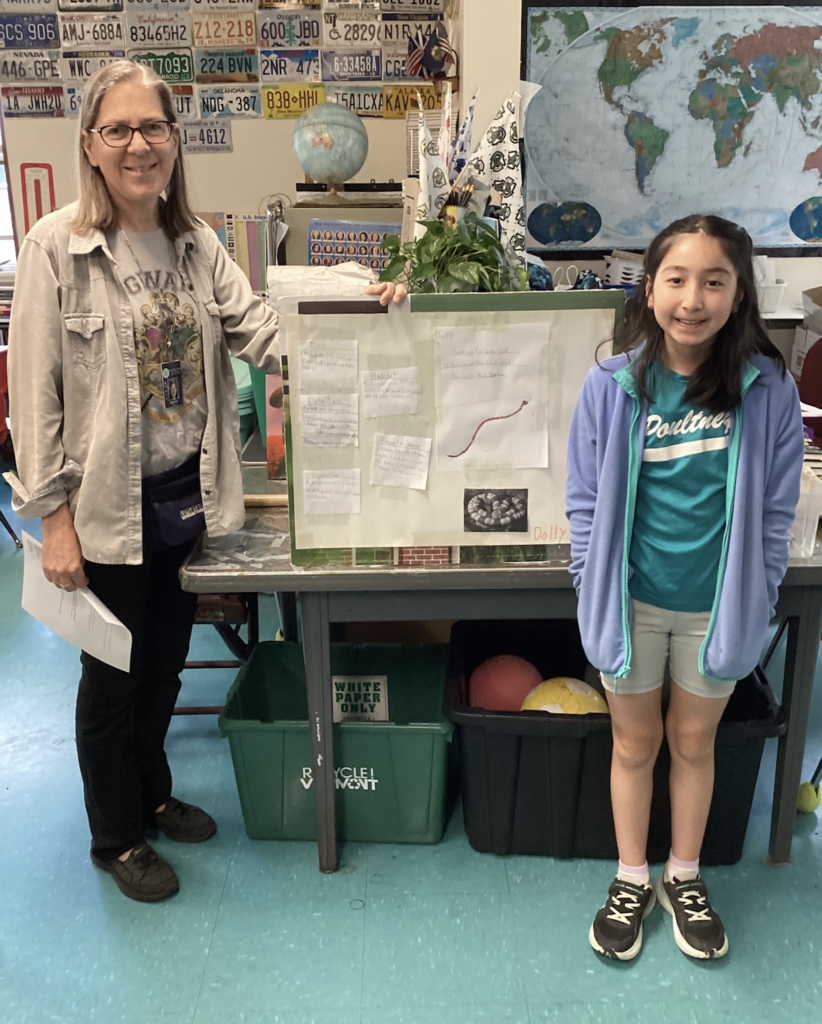 A student stands with a teacher beside a wildlife research display board featuring drawings, facts, and photographs about a snake named “Dolly.” The classroom background includes maps, globes, and educational materials.