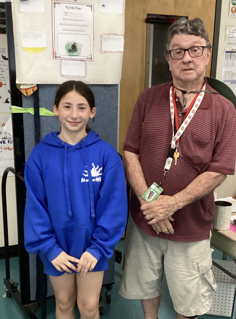 A student wearing a blue hoodie poses with a guest presenter in front of a wildlife research project display featuring a haiku poem, student writing, and a hand-drawn snail illustration.