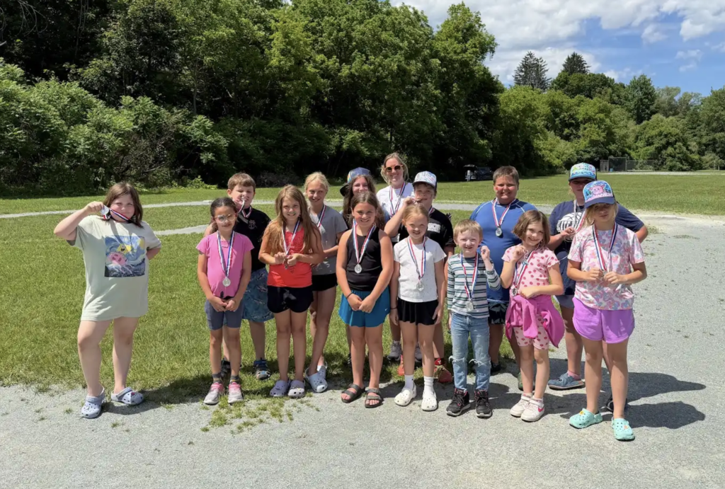 Students wearing medals gather with a teacher on a grassy field after an outdoor event. Several students proudly display their medals while posing for a group photo in the sunshine.