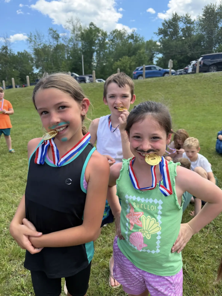 Three students proudly bite their gold medals while posing on a grassy field after an outdoor competition. Two students have playful marker mustaches and face paint as they celebrate their awards.
